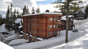 A multi-story wooden building with a snow-covered roof, surrounded by trees and a snowy landscape. - Black Diamond 3 Bedroom Condo (Whitefish)