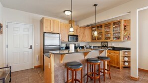 A modern kitchen with wooden cabinets, a stainless steel refrigerator, a microwave, and a bar area with stools. - Black Diamond 3 Bedroom Condo (Whitefish)