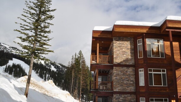 A multi-story log cabin with a stone facade, wooden balconies, and a snowy mountain backdrop. - Black Diamond 3 Bedroom Condo (Whitefish)