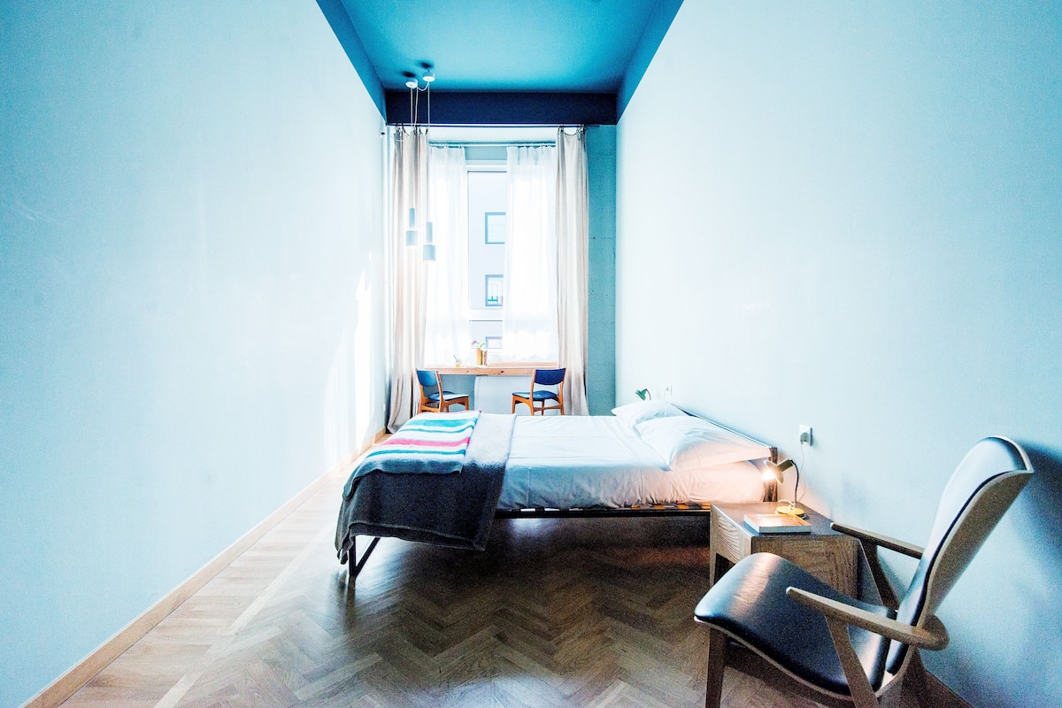 A bedroom with a bed, bedside table, chair, and a desk with a lamp.
