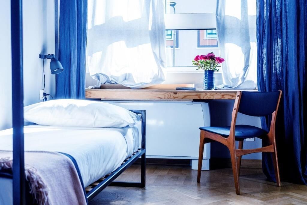 Un dormitorio con cama, escritorio, silla, refrigerador pequeño y ventana con cortinas.