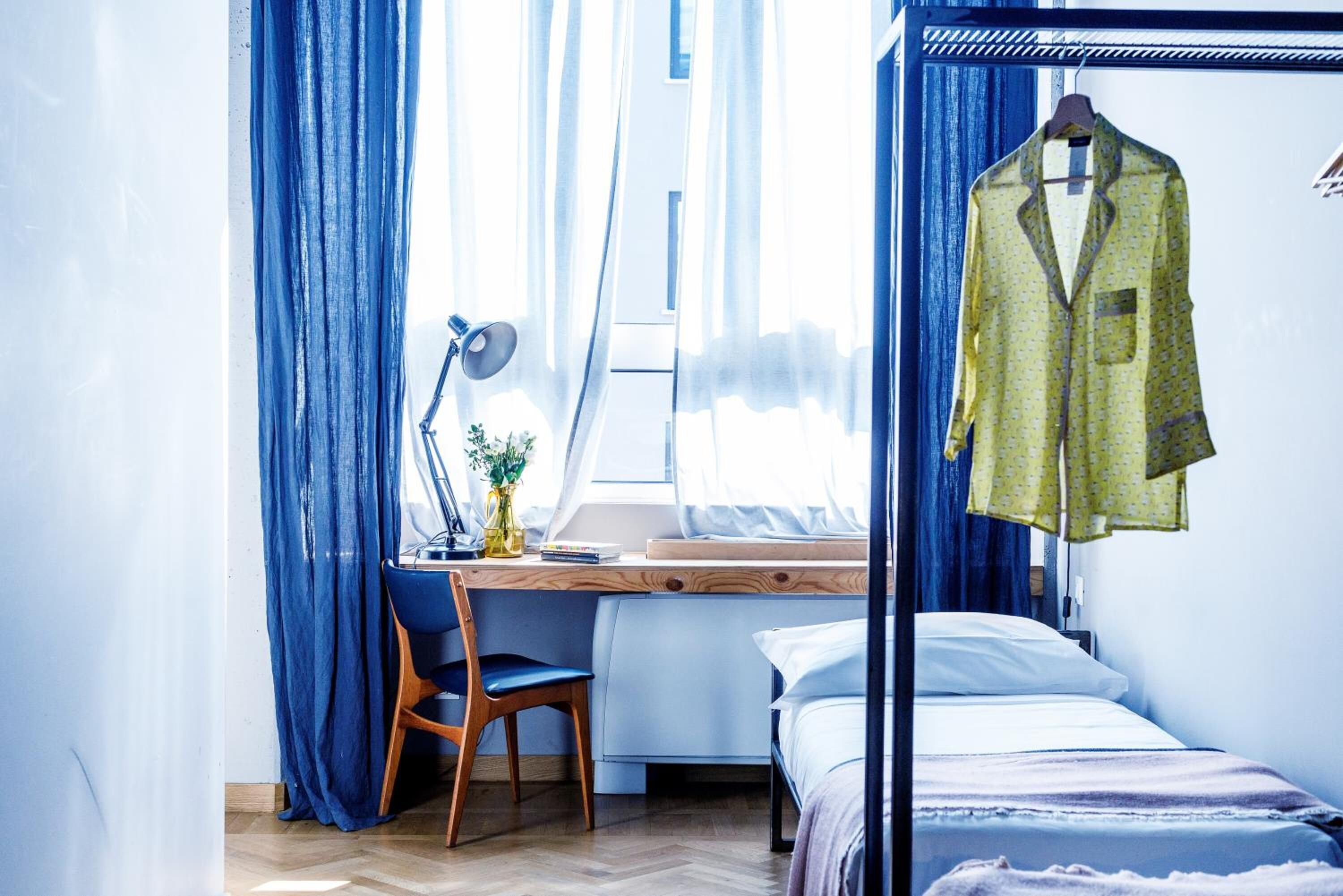 A bedroom with a bed, a desk with a chair, a hanging garment, and a window with curtains.
