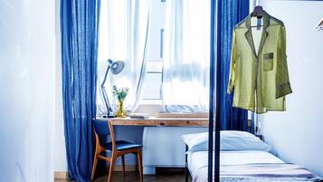 A bedroom with a bed, a desk with a chair, a hanging garment, and a window with curtains.