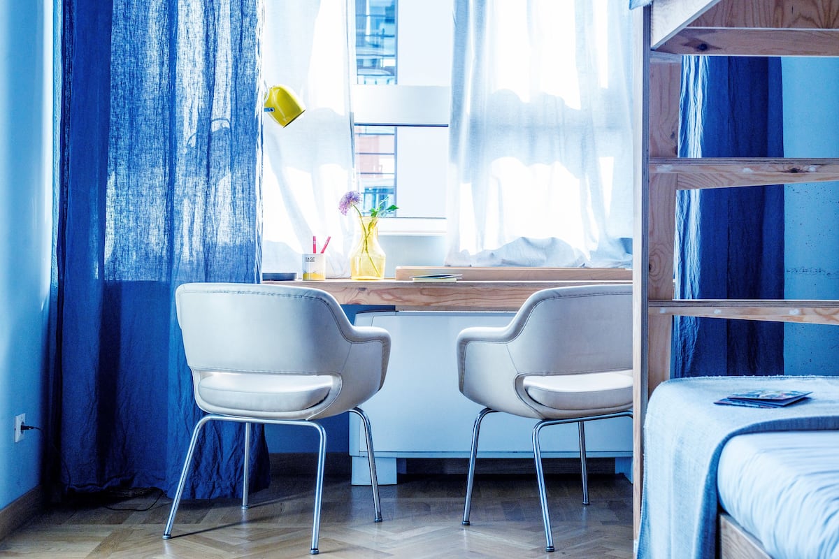 A room with a bunk bed, two white chairs, a desk with a lamp, and a window with curtains.