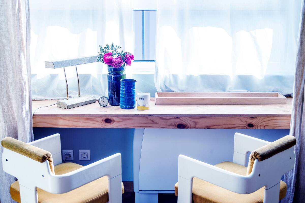 A room with a wooden desk, two white chairs with brown cushions, a vase with flowers, a clock, and a window with sheer curtains.