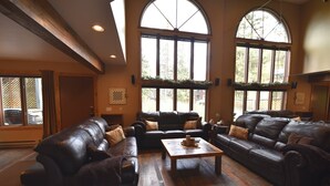 A spacious living room with leather sofas, a wooden coffee table, and large windows offering a view of the outdoors. - Whitefish Mountain 7 Bedroom Townhouse (Whitefish)