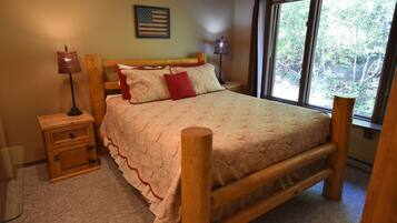 A wooden bed with a patterned bedspread and pillows, a nightstand with a lamp, and a window with a view of trees.