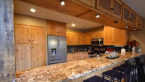 A kitchen with wooden cabinets, stainless steel appliances, granite countertops, and bar stools. - Whitefish Mountain 7 Bedroom Townhouse (Whitefish)