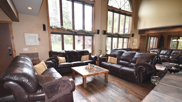 A spacious living room with leather sofas, a wooden coffee table, and large windows offering a view of the outdoors. - Whitefish Mountain 7 Bedroom Townhouse (Whitefish)