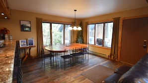A dining area with a wooden table and chairs, a chandelier, and large windows with curtains. - Whitefish Mountain 7 Bedroom Townhouse (Whitefish)