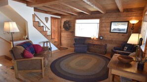 A cozy log cabin interior with a wooden staircase, a fireplace, a round rug, and a dining area with a table and chairs.
