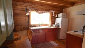 A compact kitchen with wooden walls, a window with plaid curtains, a sink, a refrigerator, and a stove.