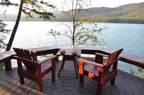 A wooden deck with Adirondack chairs and a table, overlooking a calm lake with forested hills in the background. - Ashley Lake 1 Bedroom Cabin (Kalispell)