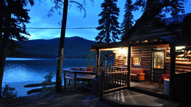 A log cabin with a deck overlooking a lake, featuring a dining area and a cozy interior with seating.