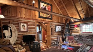A rustic cabin interior with a wooden log wall, a cozy living area with sofas and a television, and a dining table with various items.