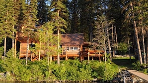 A log cabin with a deck, surrounded by dense forest and a lake.