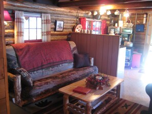 A cozy log cabin interior with a sofa, a small table with a book and a vase, and a kitchen area in the background. - Ashley Lake 1 Bedroom Cabin (Kalispell)