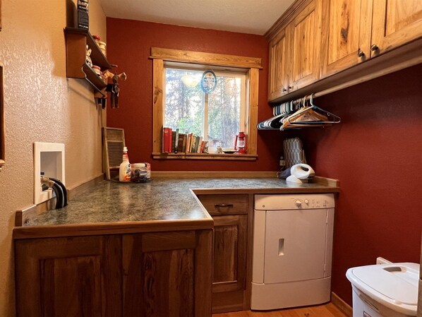 A compact kitchen with wooden cabinets, a built-in dishwasher, and a window with a view of trees. - Ashley Lake 1 Bedroom Cabin (Kalispell)