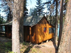A wooden cabin with a sloped roof, surrounded by tall trees. - Ashley Lake 1 Bedroom Cabin (Kalispell)