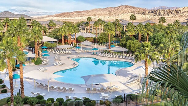 A resort with a large pool surrounded by lounge chairs and umbrellas, palm trees, and a mountainous backdrop.