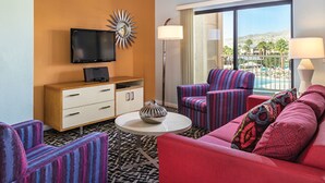 A hotel room with a large window, a flat-screen TV, a minibar, striped armchairs, a red sofa, and a patterned rug.