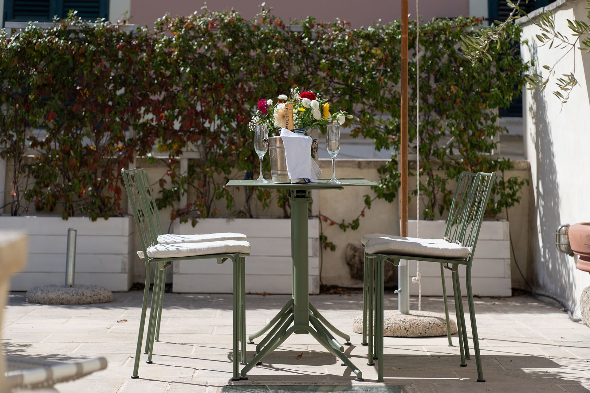 An outdoor patio with a table set for two, featuring a vase with flowers and a wine glass.
