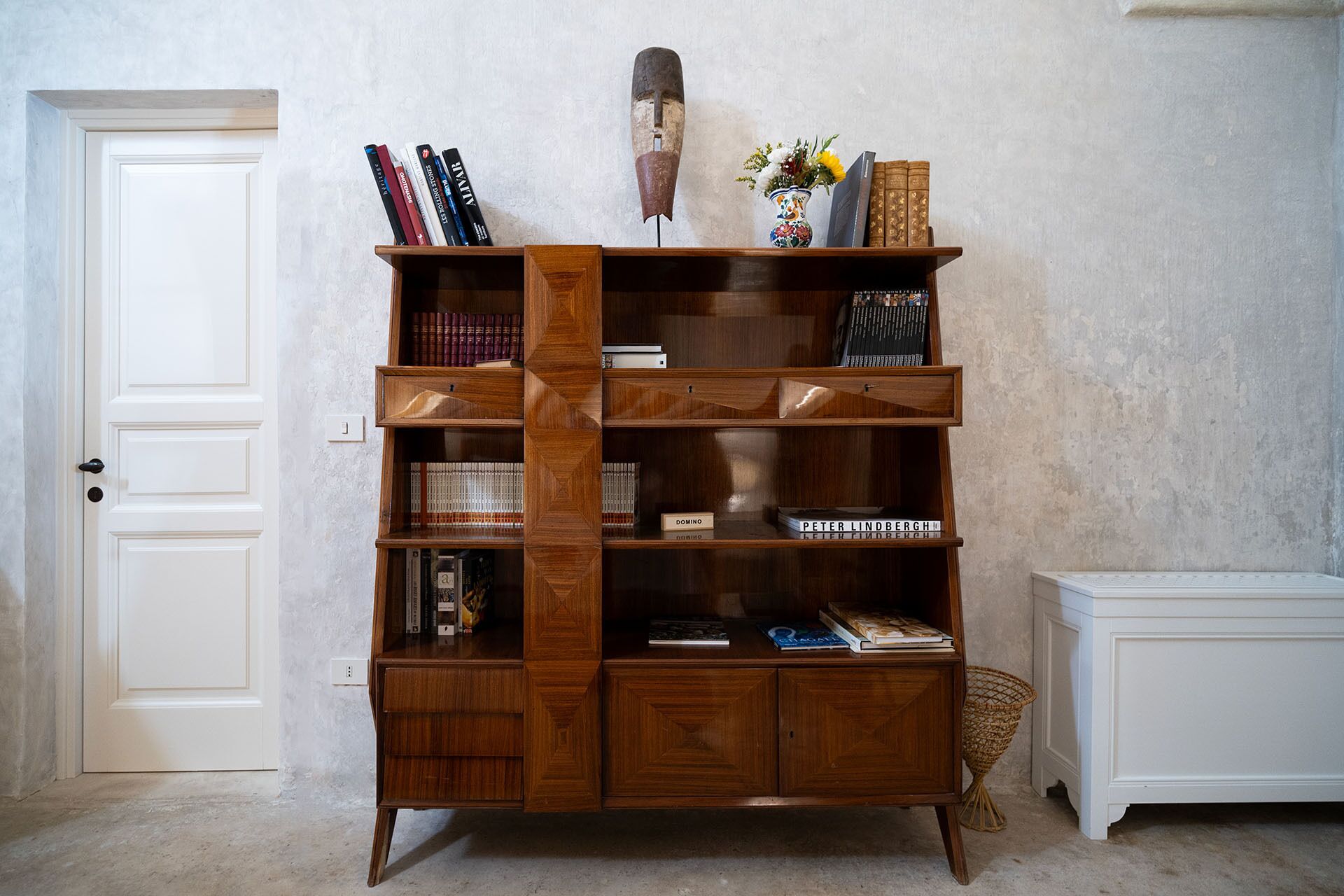 A wooden bookshelf with multiple compartments, a decorative mask, and a vase with flowers.