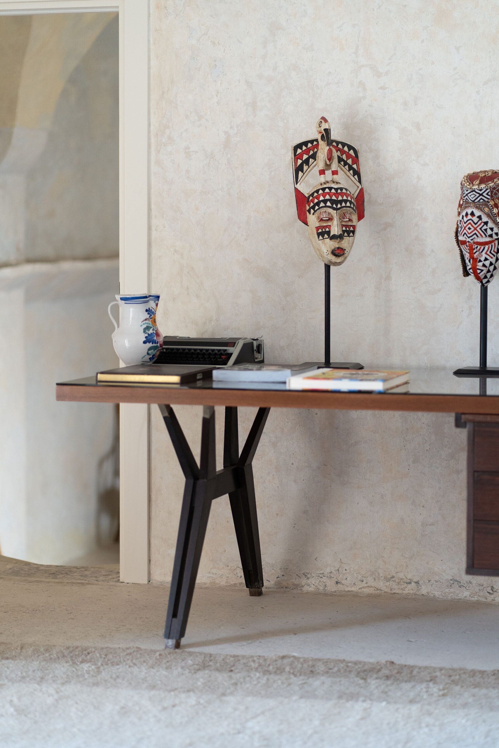 Una scrivania con un portatile, libri, una brocca in ceramica e due maschere decorative, tutto su uno sfondo di una parete strutturata.