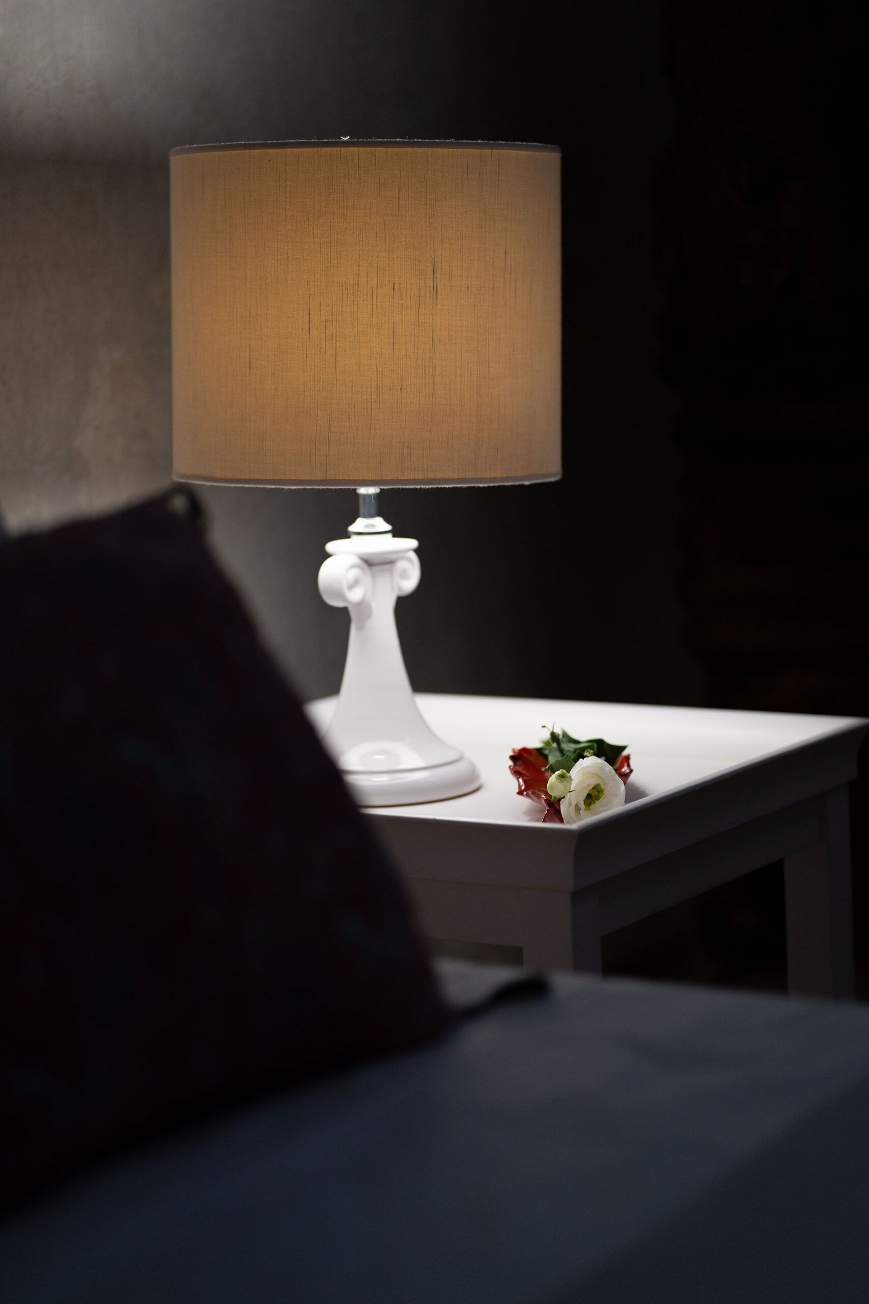 A white bedside table with a white lamp and a small bouquet of white flowers.