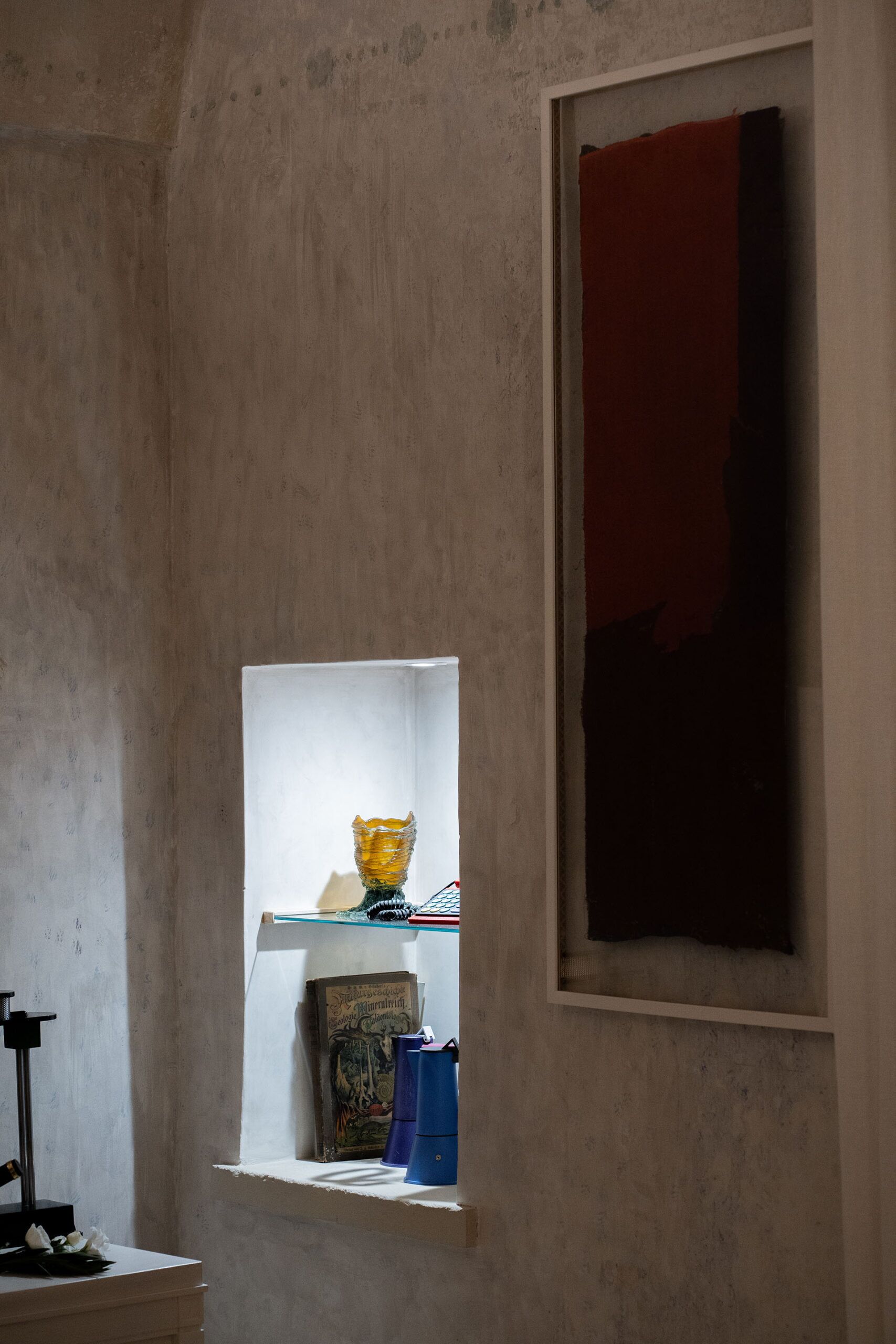 Une pièce avec une petite fenêtre, une étagère sur laquelle se trouvent un livre et un vase, et un objet rouge accroché au mur.