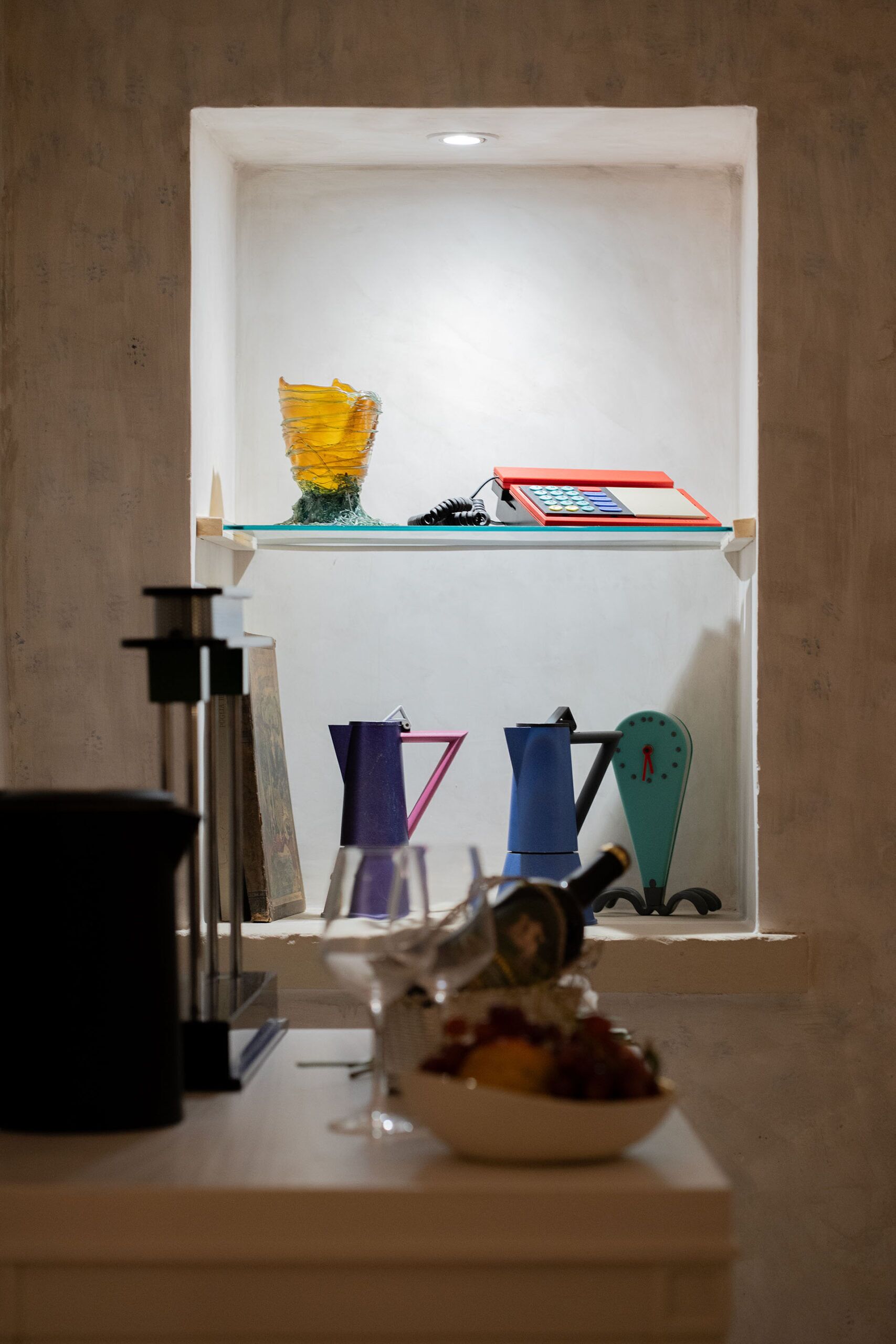 A small, well-lit room with a white shelf containing various items including a yellow object, a red box, and a blue item.