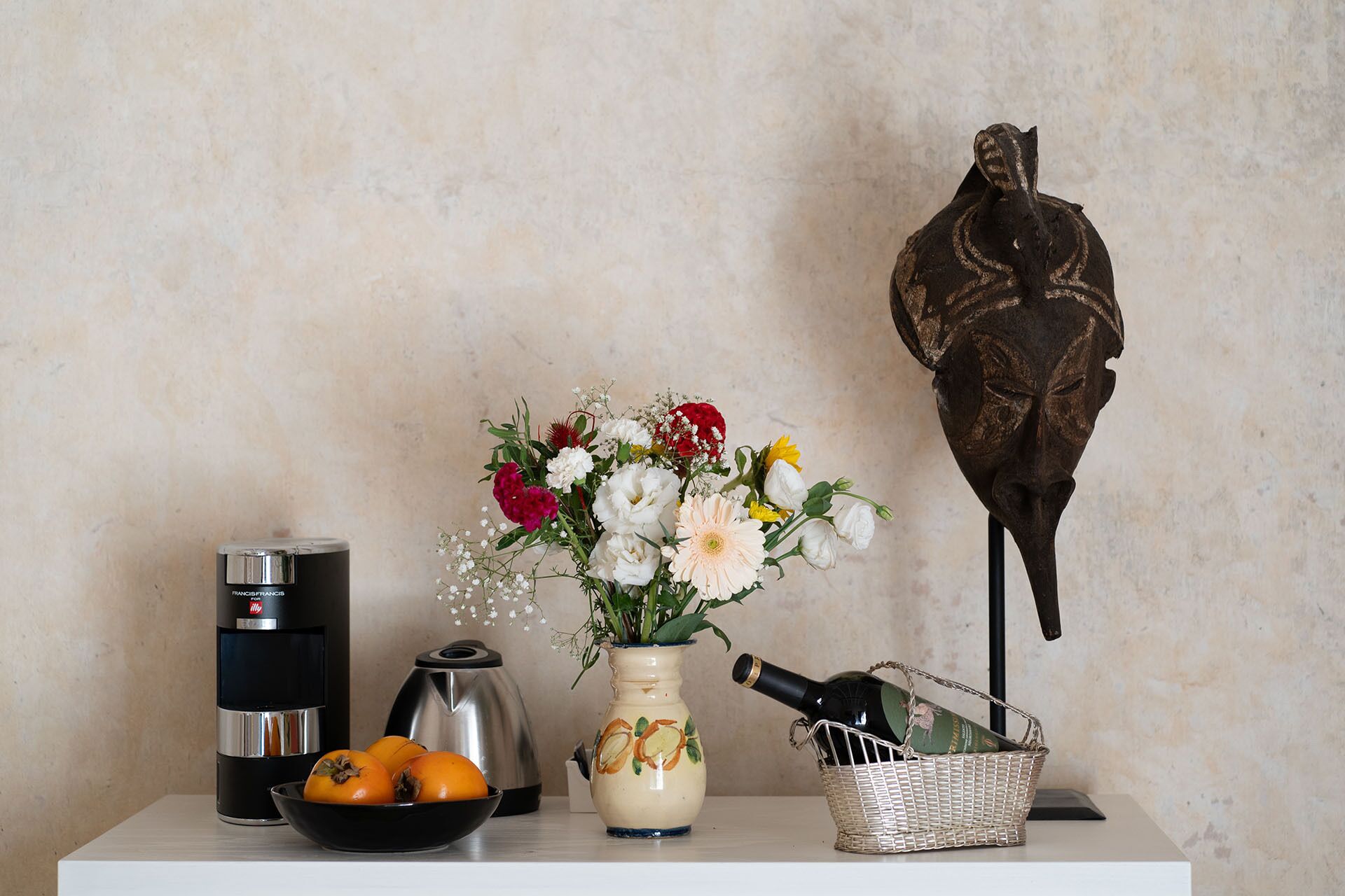 A kitchen counter with a coffee maker, a kettle, a bowl of oranges, a vase with flowers, and a wine bottle in a basket.