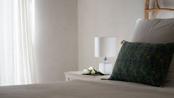Una cama bien hecha con un almohadón blanco y uno verde estampado, una mesita de noche blanca con una lámpara, y una silla de mimbre.