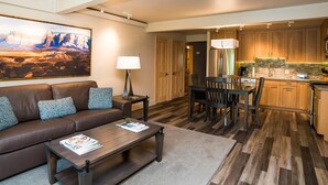 A modern living room with a brown leather sofa, a wooden coffee table, and a dining area with wooden chairs. A kitchen with wooden cabinets and stainless steel appliances is visible in the background. - Lift One - Recently renovated one bedroom with patio (Aspen)