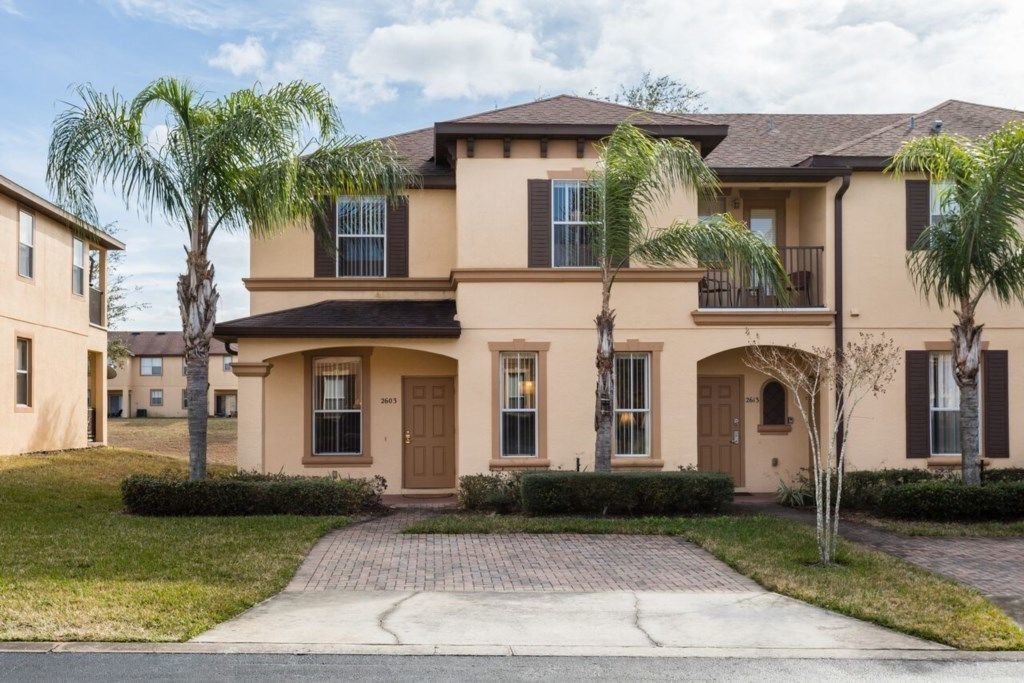 A two-story residential building with a beige facade, multiple windows, and a brick driveway.