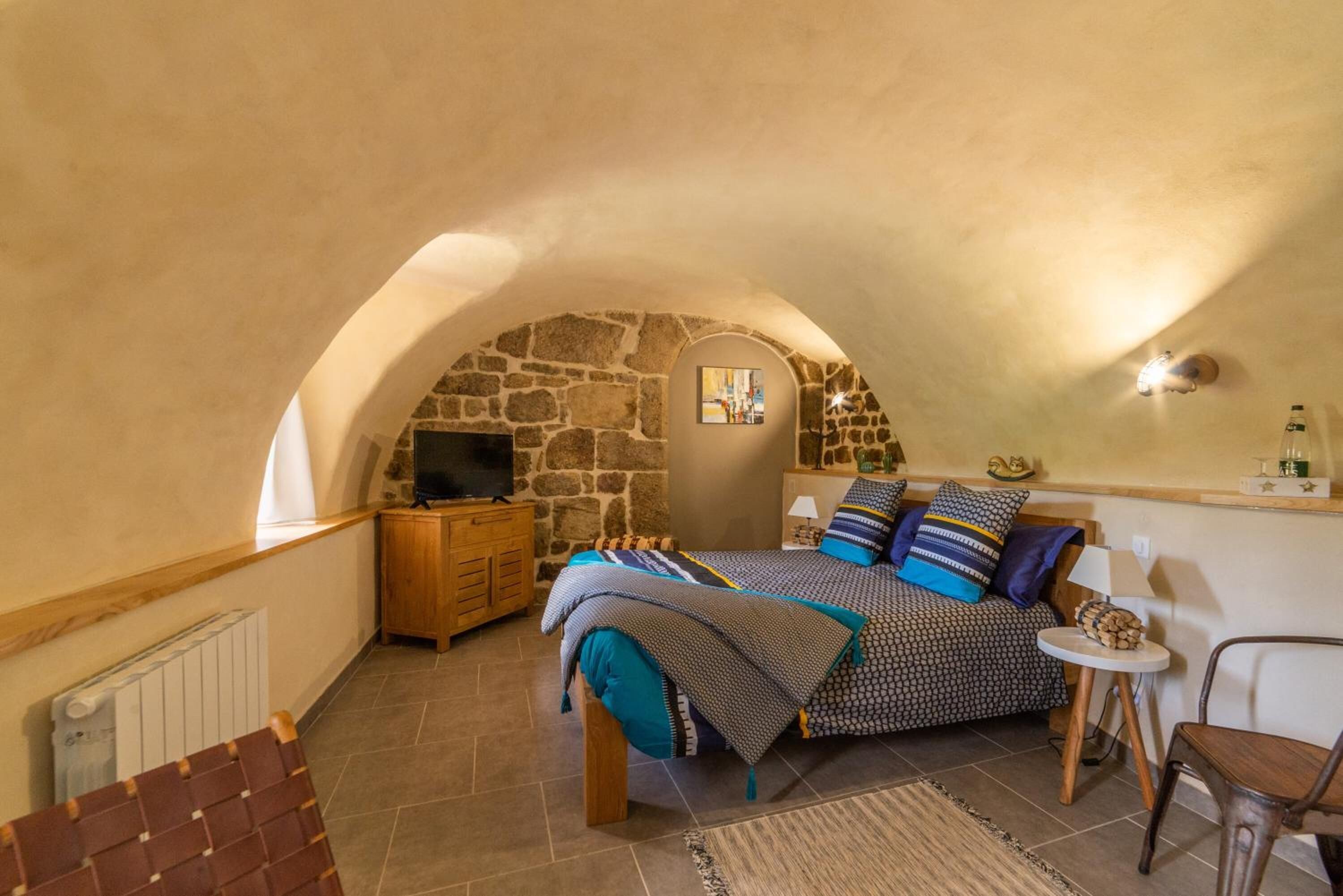 A cozy bedroom with a stone wall, a bed with patterned bedding, a small wooden dresser, a television, and a radiator.