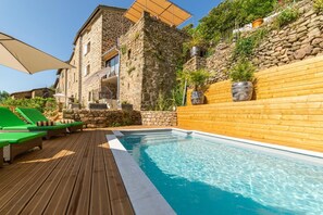 A pool area with wooden deck, lounge chairs, and a stone building with a wooden wall and potted plants. - La Calade (Chirols)