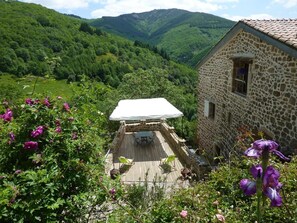 A stone building with a wooden deck and a covered seating area, surrounded by lush greenery and mountains. - La Calade (Chirols)