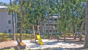 A playground with a swing set and a slide, surrounded by trees and a building in the background. - Very Desirable 1st Floor Villa at Fiddler's Cove Beach & Racquet Club (Hilton Head Island)