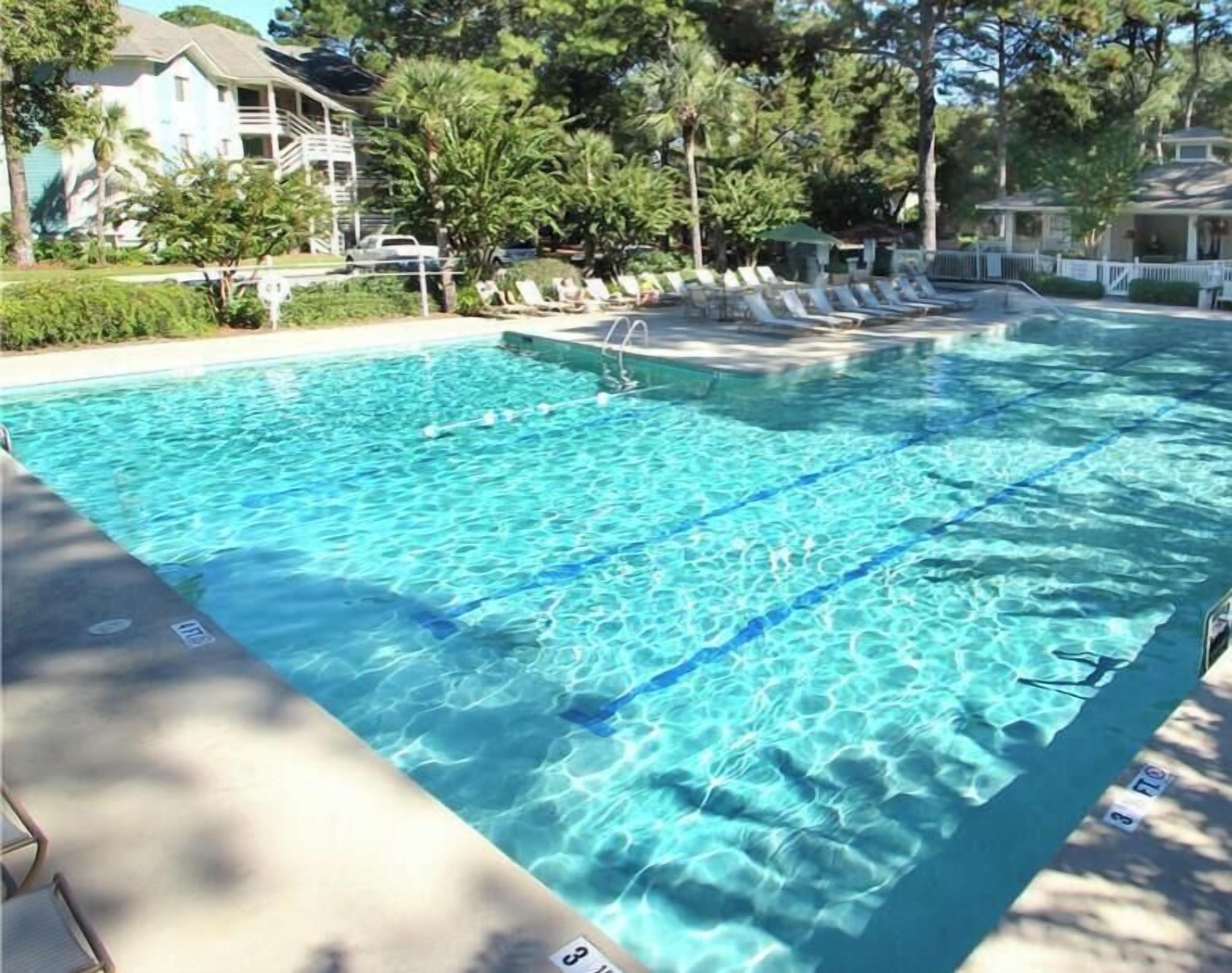 A rectangular swimming pool with clear blue water, surrounded by lounge chairs and a white fence.
