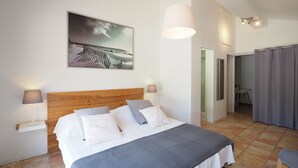 A bedroom with a wooden headboard, a large bed with white and grey bedding, a grey throw blanket, two bedside lamps, a wall-mounted picture of a beach, and a bathroom visible through an open door.