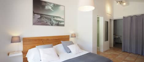 Un dormitorio con cabecera de madera, una cama grande con ropa de cama blanca y gris, una manta gris, dos lámparas de noche, un cuadro colgado en la pared con una playa, y un baño visible a través de una puerta abierta.