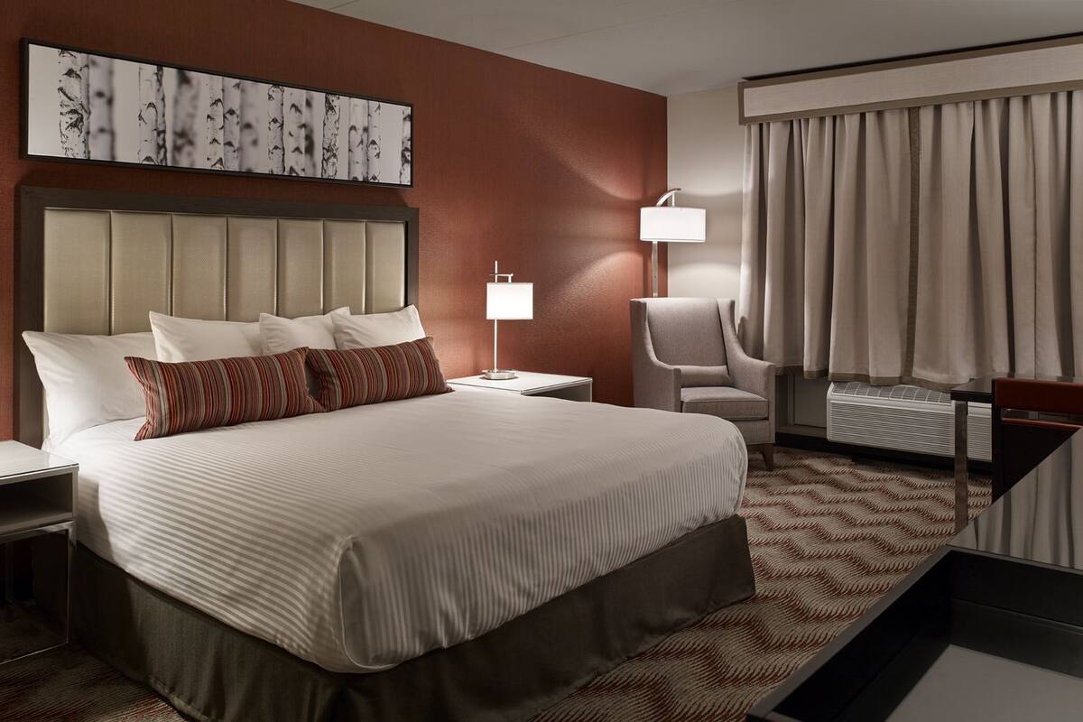 A hotel room with a large bed, a headboard, a bedside table, a chair, a lamp, a window with curtains, and a wall art piece.