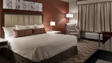 A hotel room with a large bed, a headboard, a bedside table, a chair, a lamp, a window with curtains, and a wall art piece.