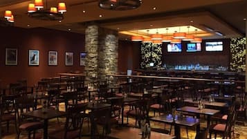 Un restaurante con varias mesas y sillas, un pilar de piedra y una zona de bar con televisores.