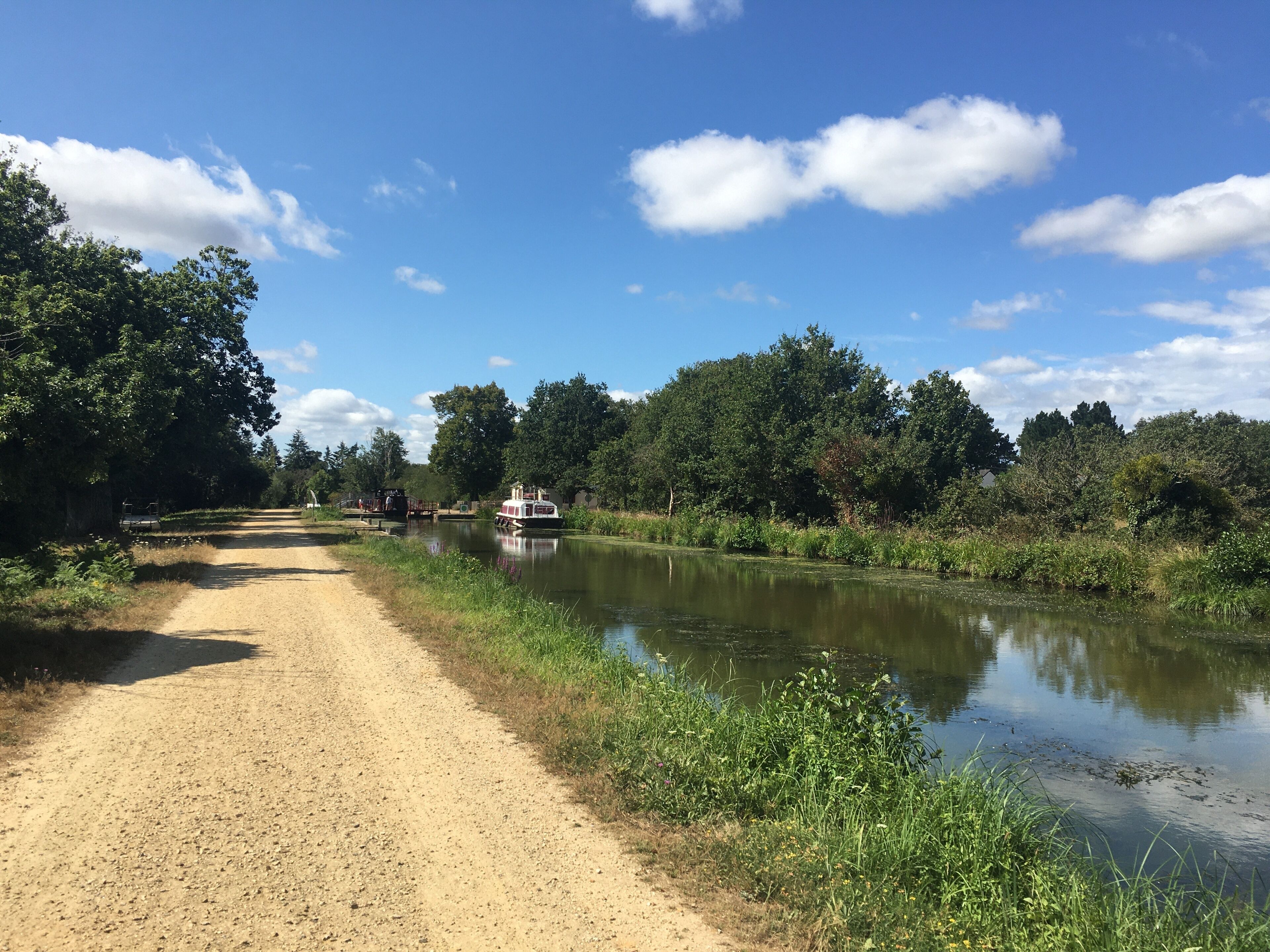Ein schmaler Feldweg entlang eines ruhigen Flusses, an dem ein Boot liegt, umgeben von dichtem Grün und Bäumen.
