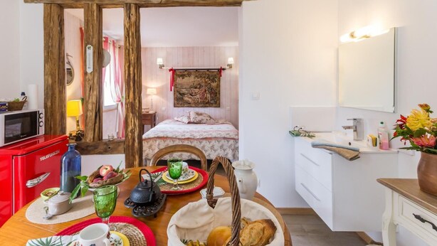 Une pièce chaleureuse avec un plafond orné de poutres en bois, un lit recouvert d’une courtepointe à motifs floraux, une table à manger avec de l’assiette et une bouilloire, et un appareil électroménager rouge.