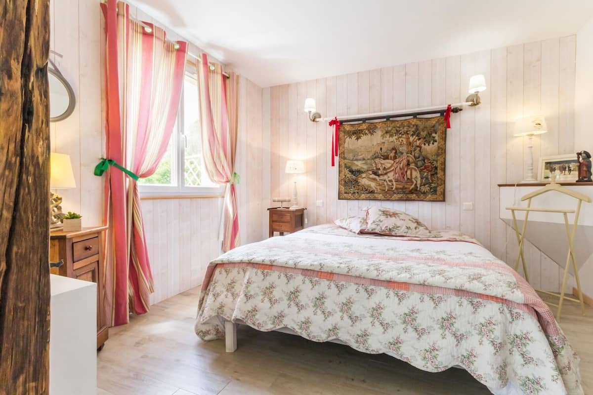 Ein Schlafzimmer mit Bett, Schreibtisch, Stuhl, einem Fenster mit Vorhängen und einem Gemälde an der Wand.