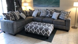 A grey sectional sofa with patterned cushions and a matching ottoman, a painting of a wave on the wall, and two floor lamps.
