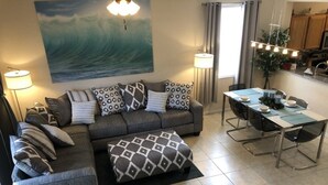 A living room with a grey sofa, patterned cushions, a dining table with chairs, a lamp, and a large painting of a wave.